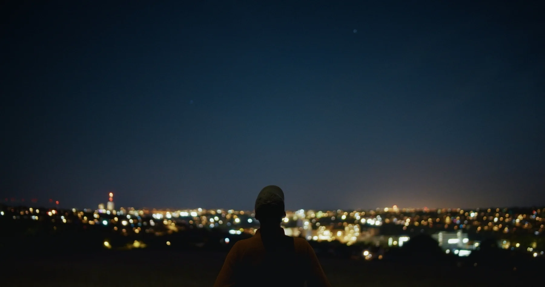 A silhouetted figure looking out over a brightly lit city at dusk